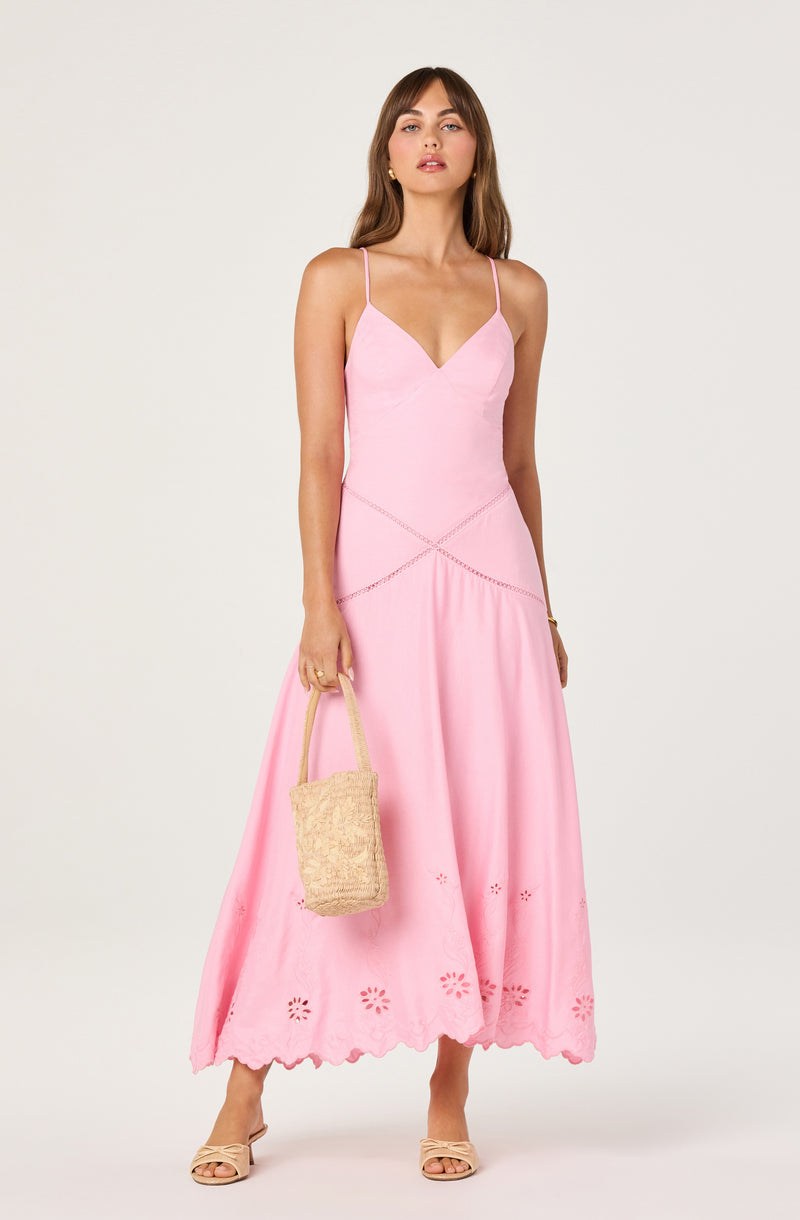 Woman wearing a pink summer dress with spaghetti straps, embroidery, and scalloped hem, holding a woven bag.