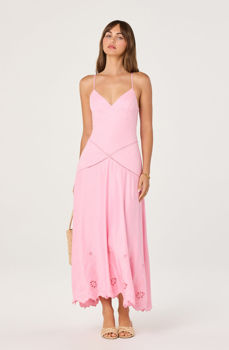 Woman in a pink spaghetti strap maxi dress with embroidered details at the hem, holding a woven bag.