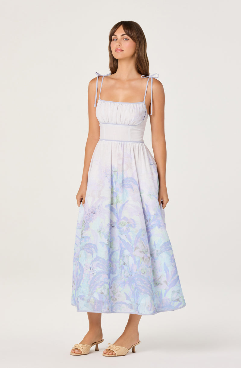 Woman in a white dress with pastel floral pattern, spaghetti straps with bows, and beige heels.