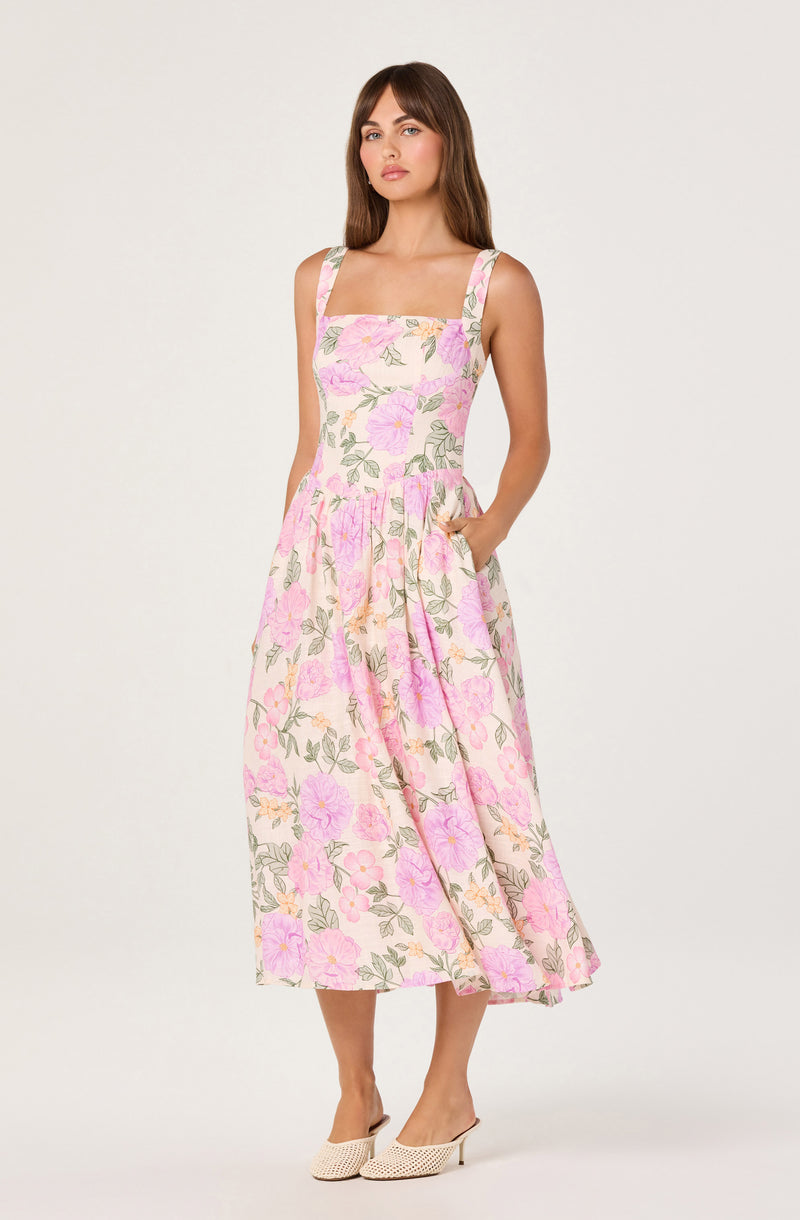 Woman wearing a pink and green floral sleeveless maxi dress with pockets and beige slip-on shoes.