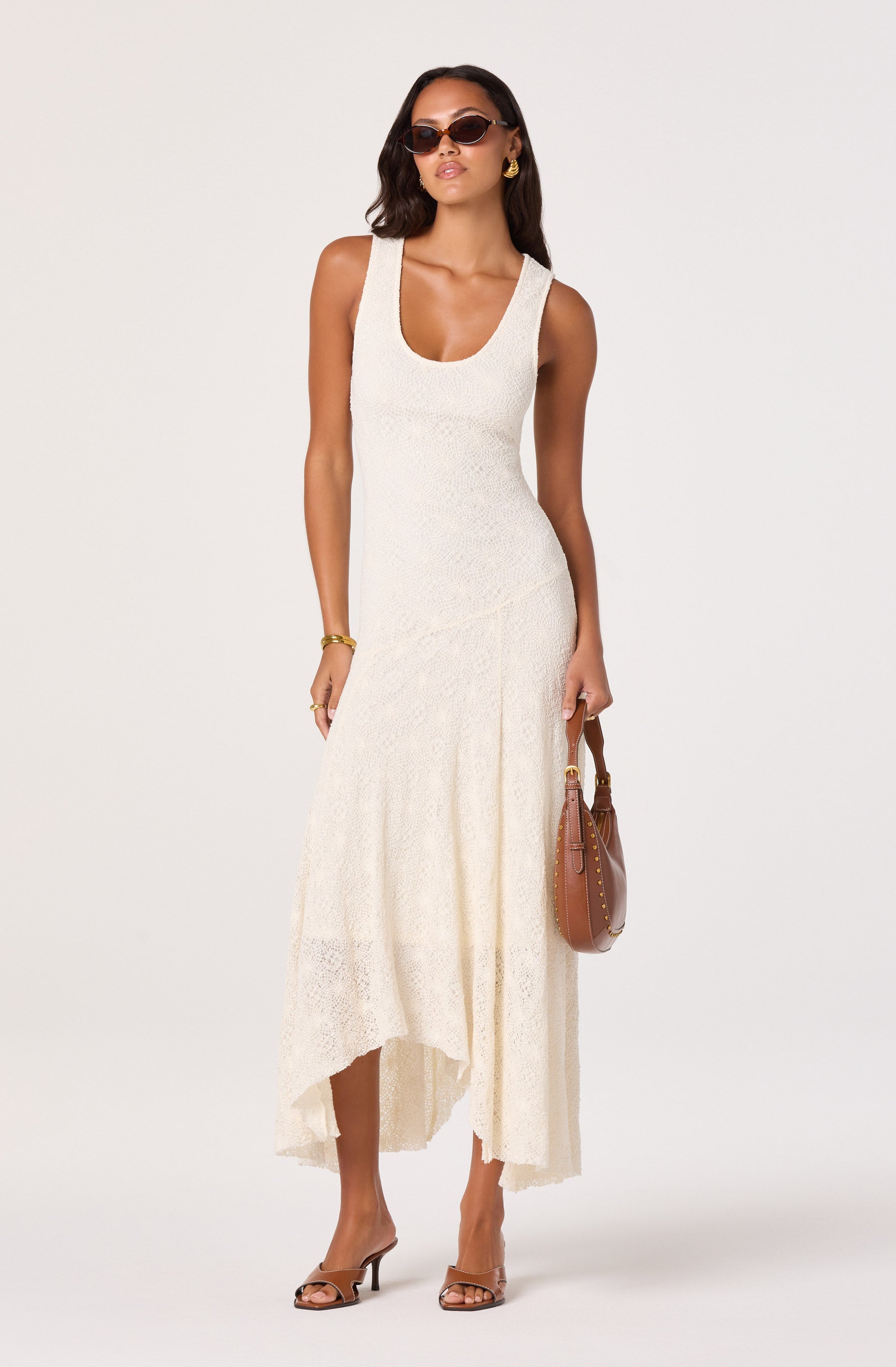 Woman in a sleeveless, cream-colored, lace maxi dress with a high-low hem, paired with tan heels, sunglasses, and gold accessories.