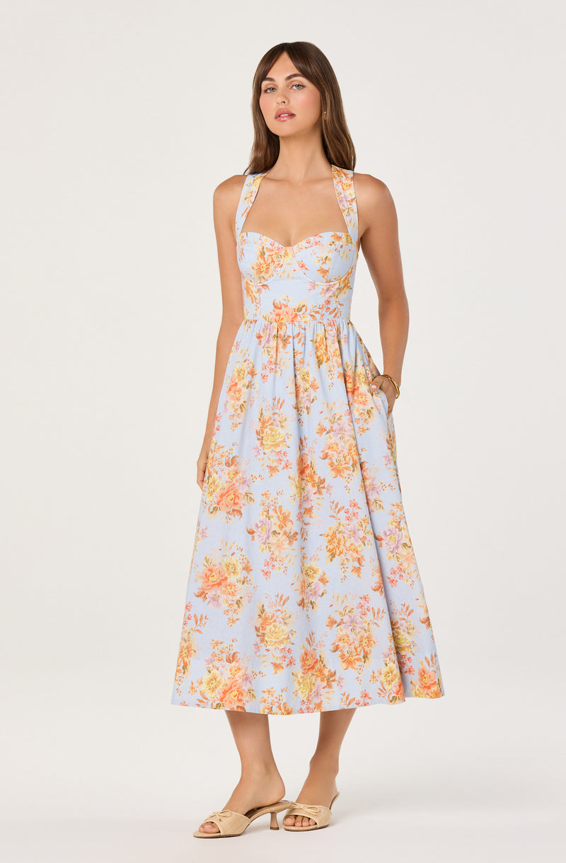 Woman wearing a sleeveless, floral print, full-length dress with wide straps and pockets.