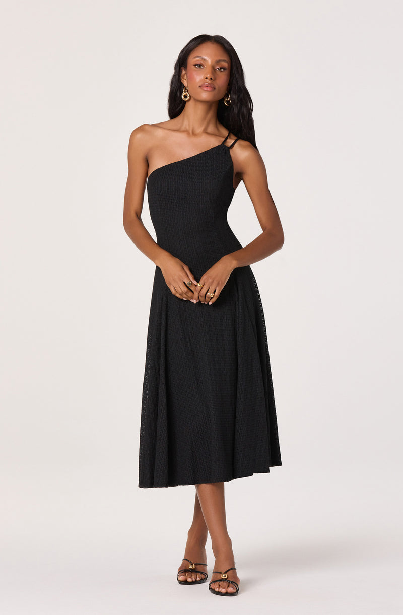Woman in a black one-shoulder midi dress with a slit, wearing gold hoop earrings and black strappy heels.