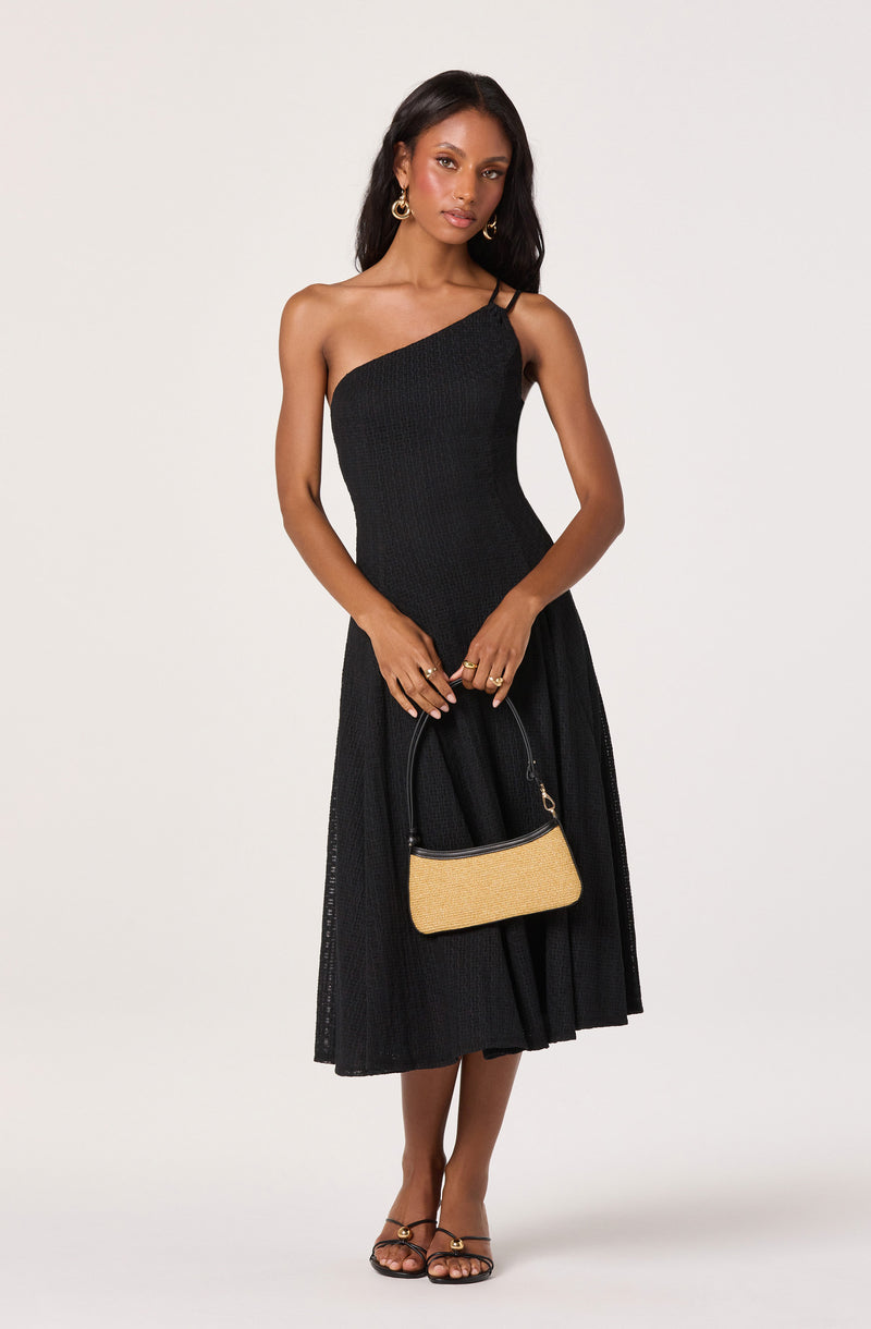 Woman wearing a black one-shoulder dress with thin shoulder straps, holding a beige handbag.