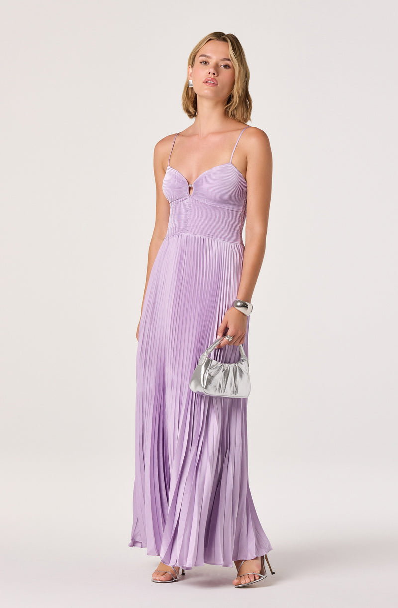 Woman in a long, pleated lavender dress with spaghetti straps, holding a silver handbag.