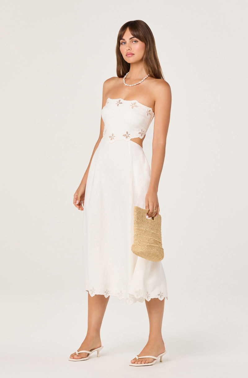 White strapless dress with floral cutouts and scalloped hem, paired with beige heels and a woven bag.