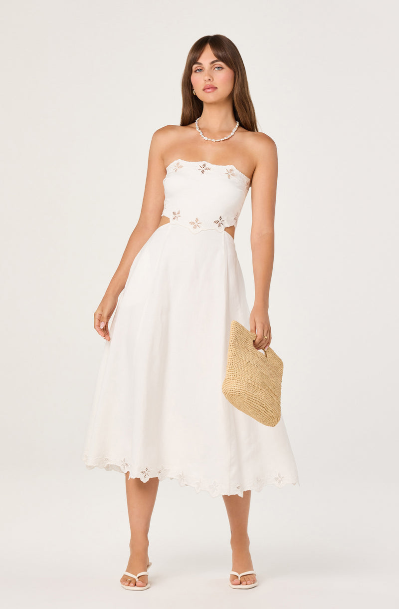 Woman in white strapless dress with floral cutouts, holding a straw handbag, and wearing white heels.