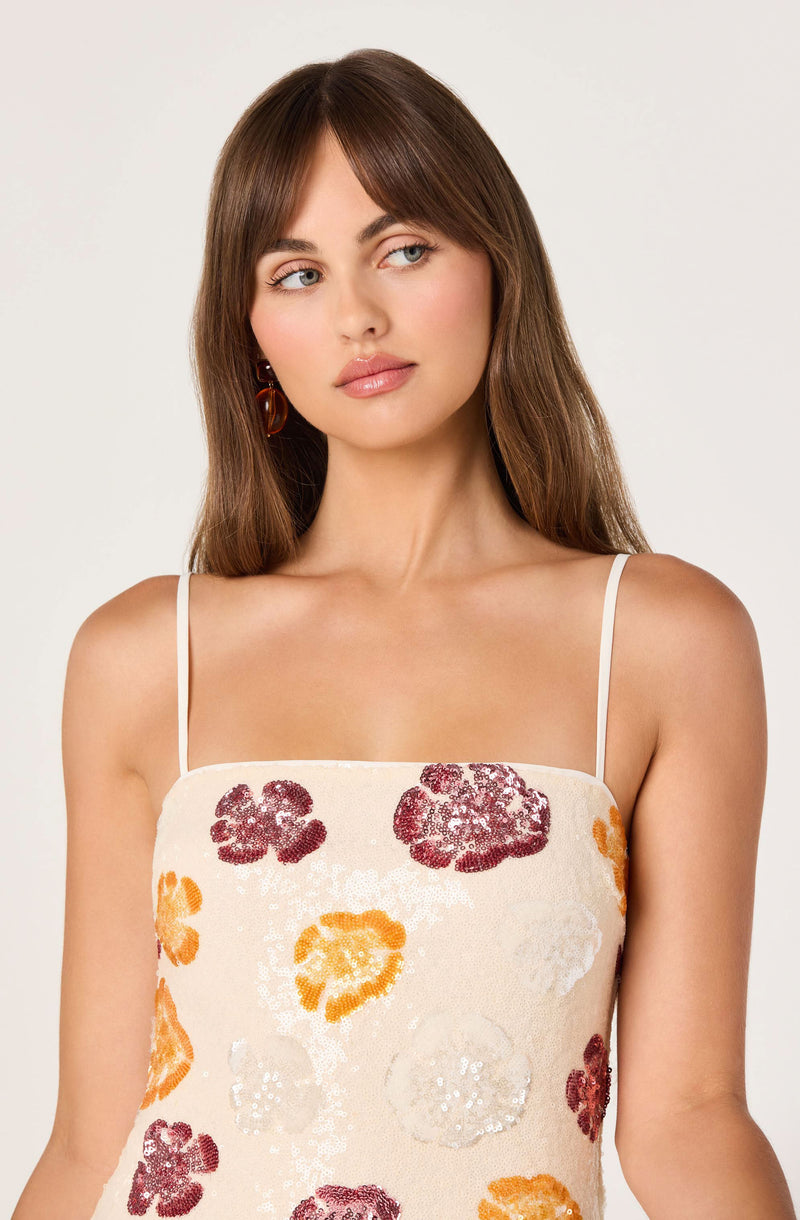Woman in a cream-colored dress with orange, red, and white floral sequin patterns and thin shoulder straps.