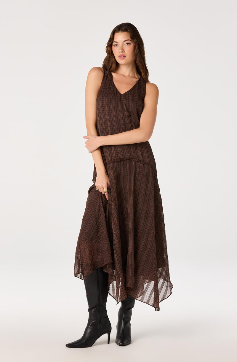 Woman wearing a brown sleeveless dress with a V-neck and a sheer, asymmetrical skirt, paired with black high-heeled boots.