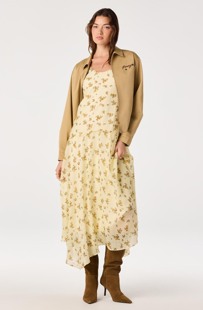 Model wearing a beige jacket over a cream dress with gold floral patterns, paired with brown knee-high boots.