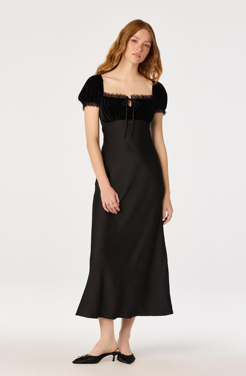 Woman in black dress with puffed short sleeves, lace details, and flat shoes with bows.