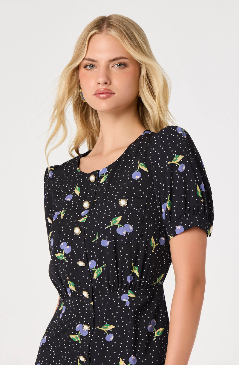 Woman wearing a black dress with a cherry and leaf pattern, puffed short sleeves, and pearl button details.