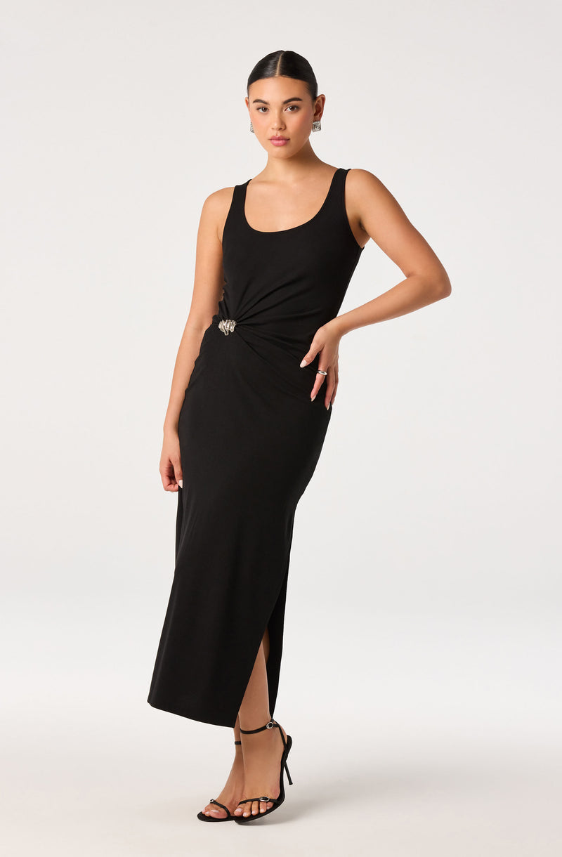 Woman in a long black sleeveless dress with side slit and decorative pin, wearing black strappy heels.