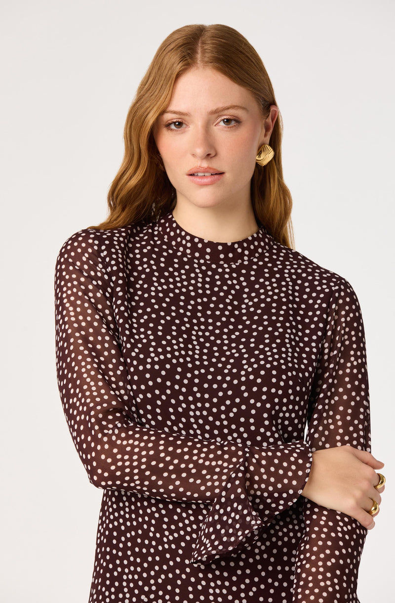 Woman wearing a dark brown long-sleeve dress with white polka dots, and gold earrings.