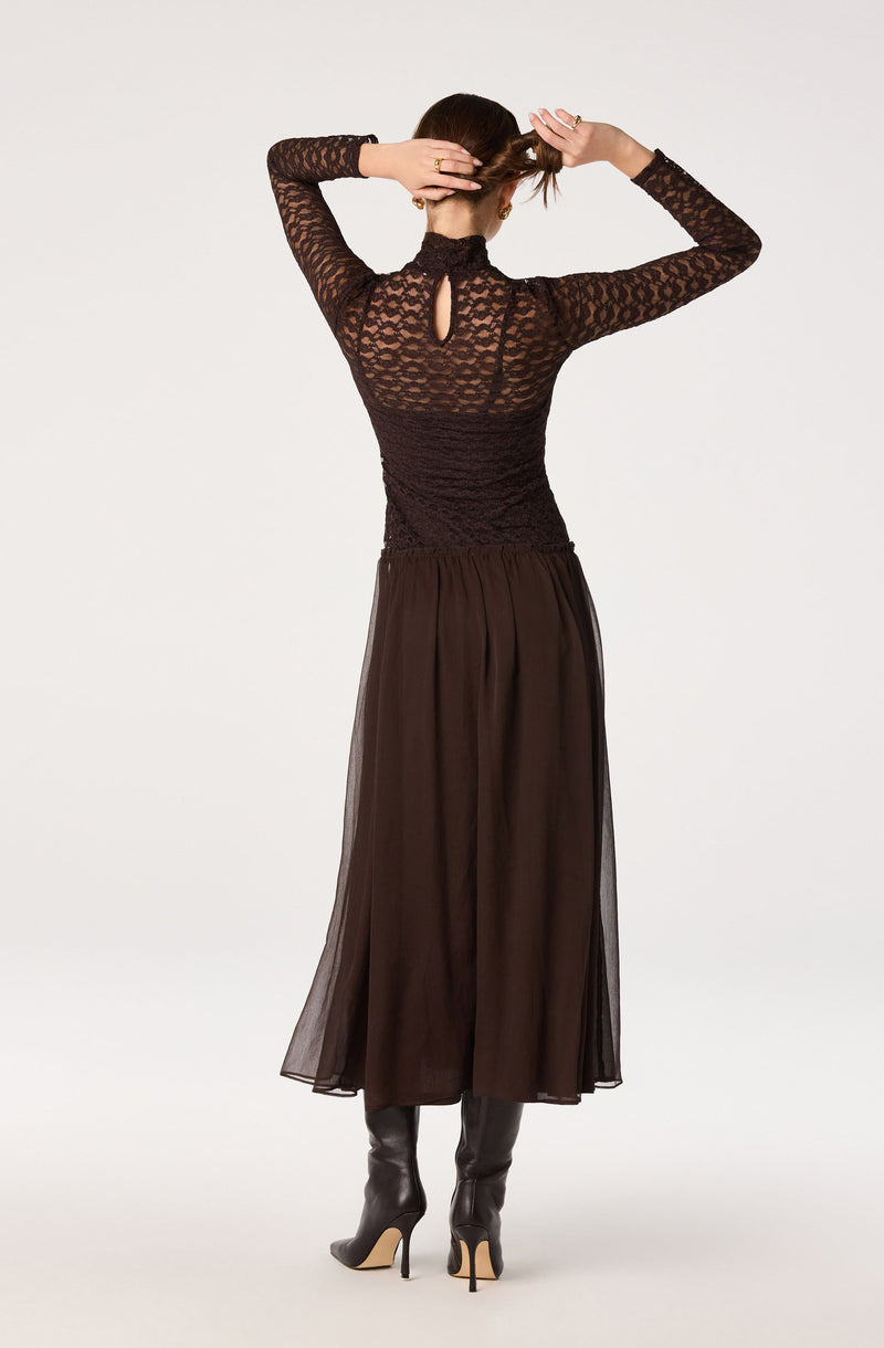 Woman in a long black lace top with a keyhole back, paired with a sheer black skirt and tall black boots.
