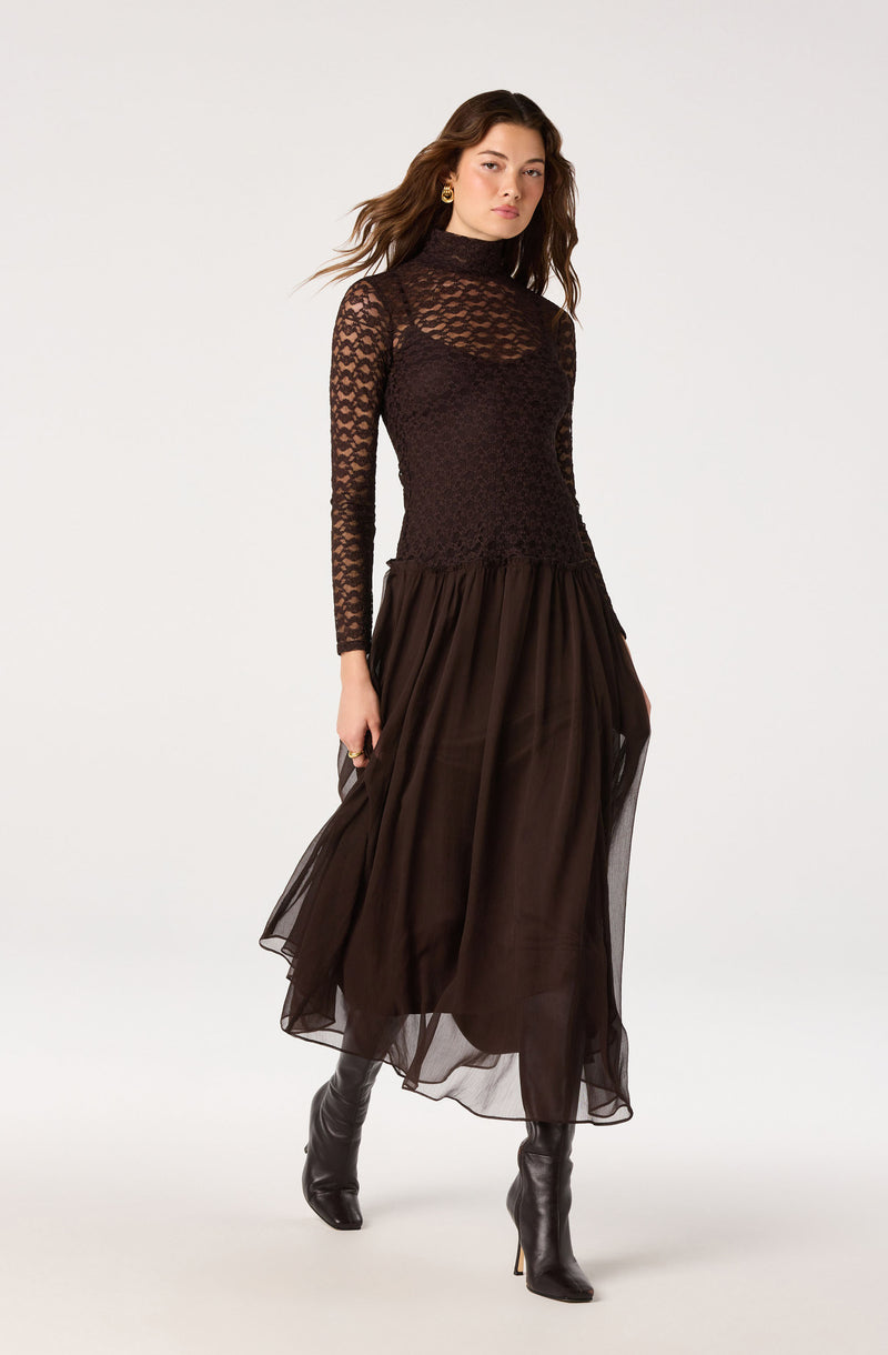 Woman in black lace turtleneck top and flowing brown skirt with knee-high black boots.