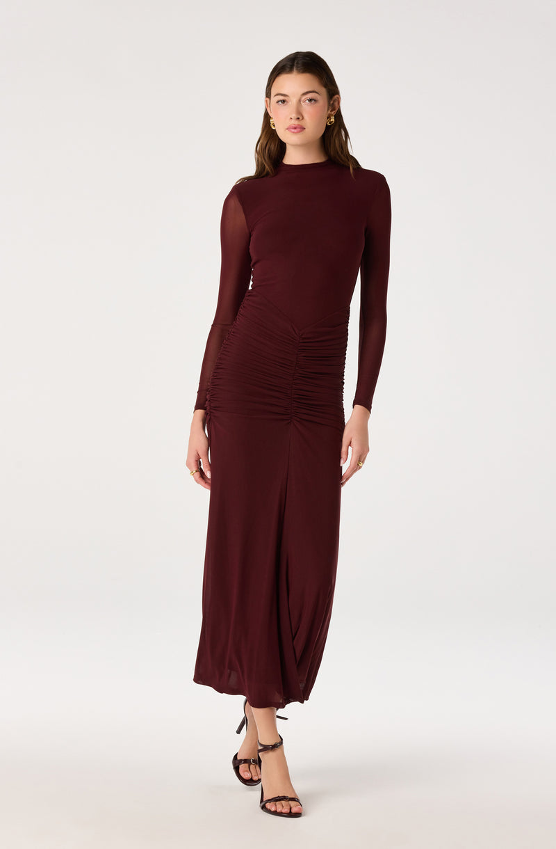 Woman in a long, ruched maroon dress with sheer long sleeves and black high-heeled sandals.