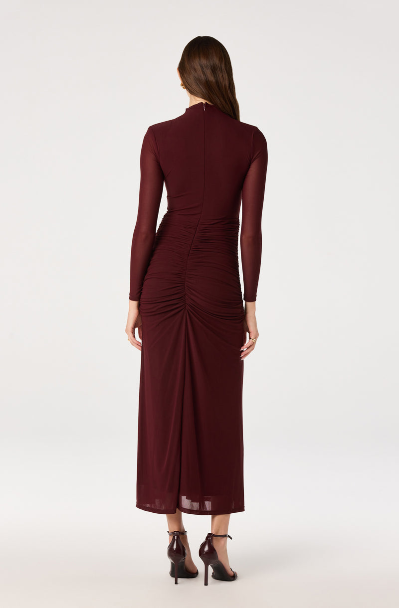 Woman in a long, burgundy, fitted dress with ruching and sheer long sleeves.