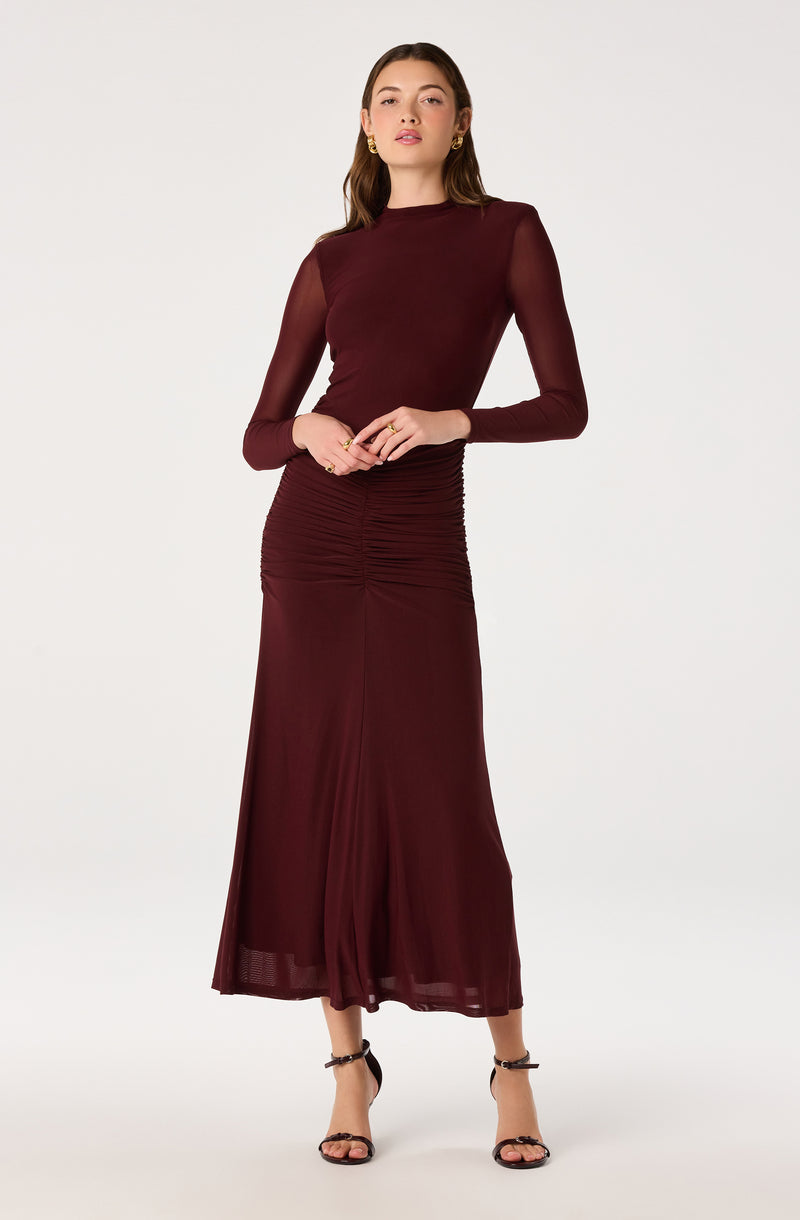 Woman wearing a long, burgundy dress with sheer long sleeves and ruched detail at the waist.
