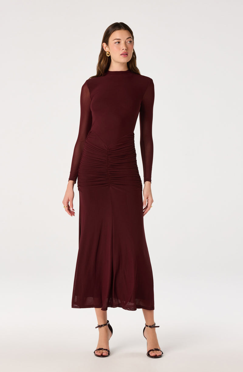 Woman wearing a long, burgundy, sheer, long-sleeve dress with ruched detail at the waist and black strappy heels.