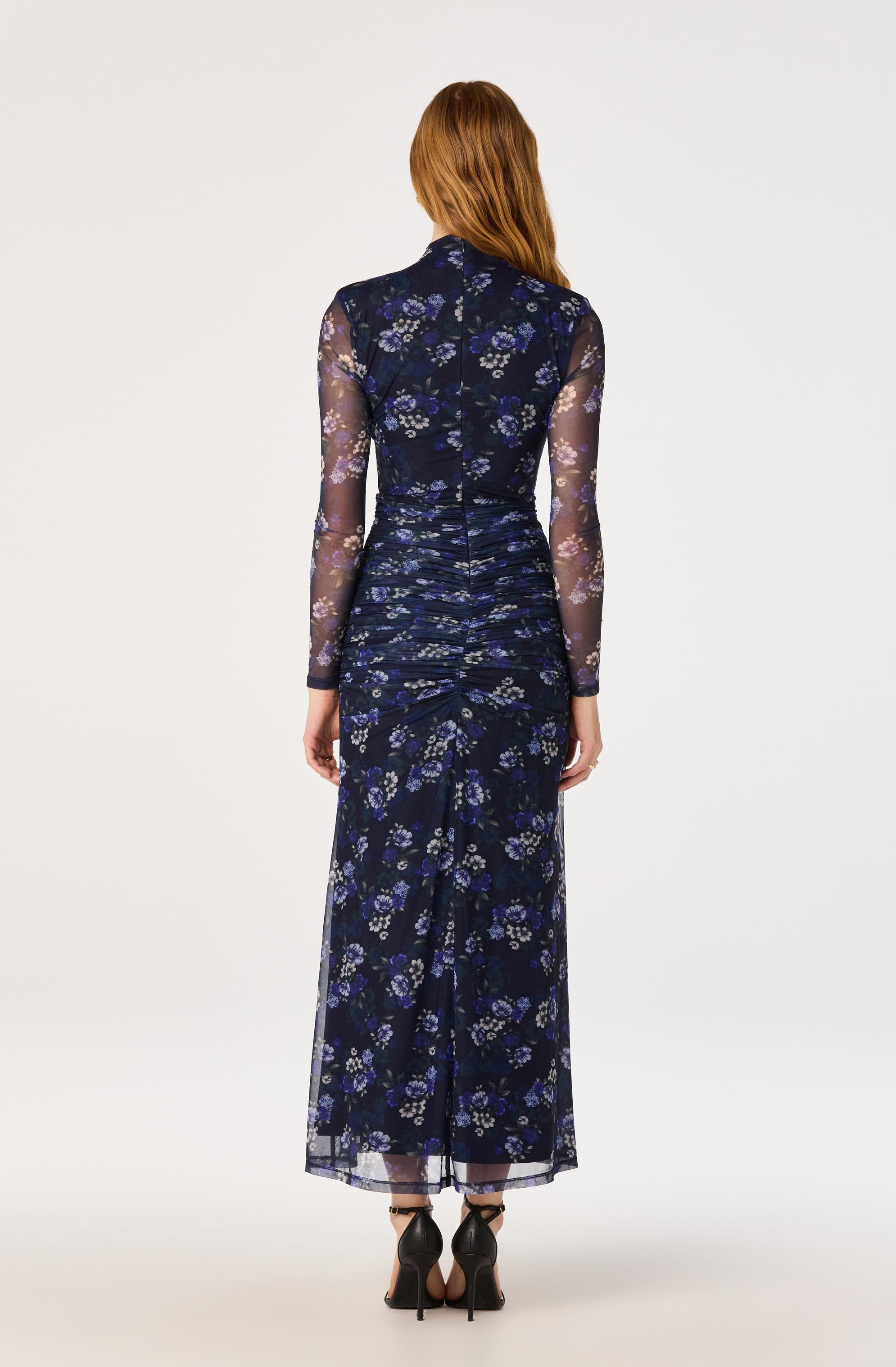 Model wears a long, navy floral dress with sheer long sleeves and ruched fabric.