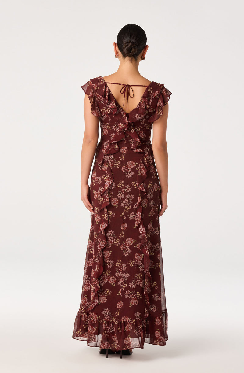 Back view of a woman wearing a long, burgundy floral dress with ruffles and a tie-back detail.