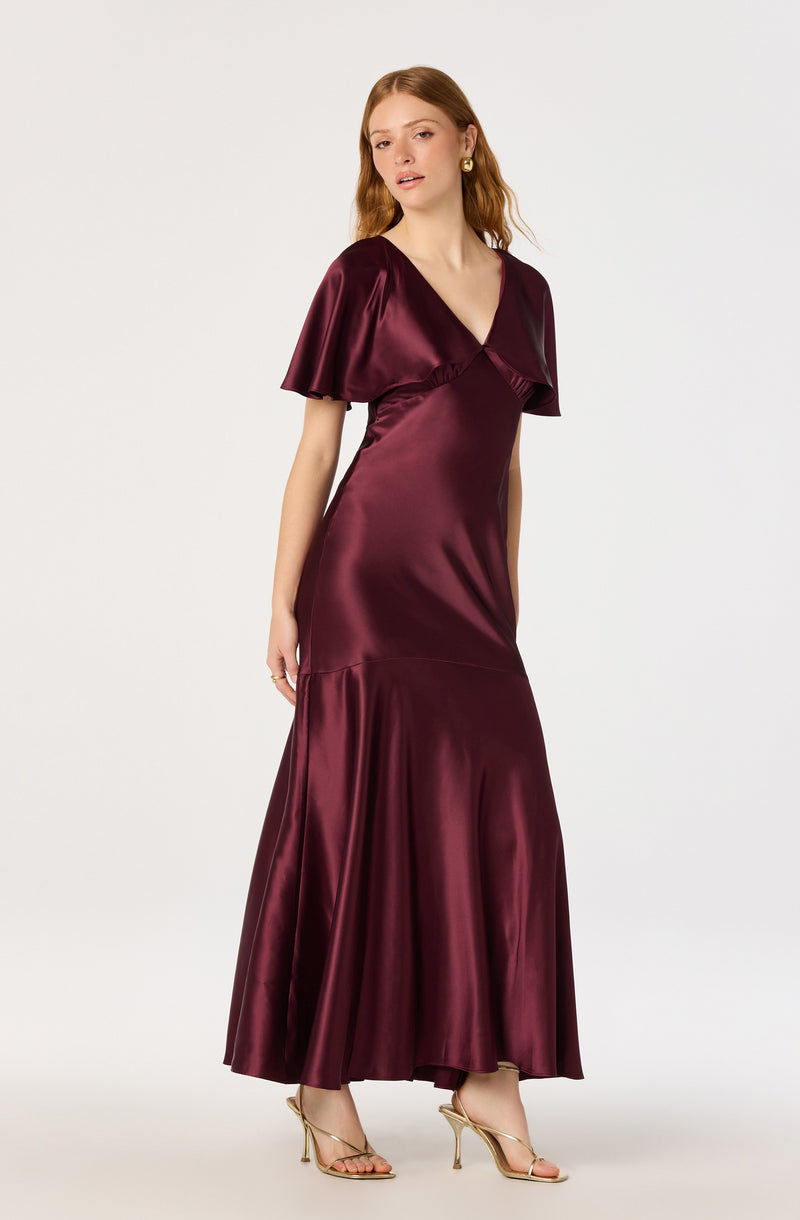 Woman in a satin, deep burgundy, floor-length dress with ruffle sleeves and gold heels.
