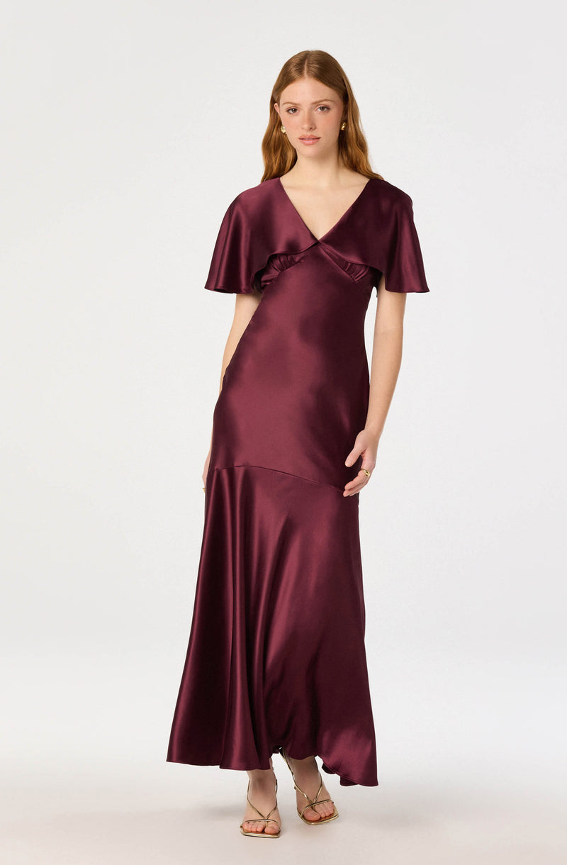Woman wearing a long, burgundy satin dress with short, cape-like sleeves and a V-neck.
