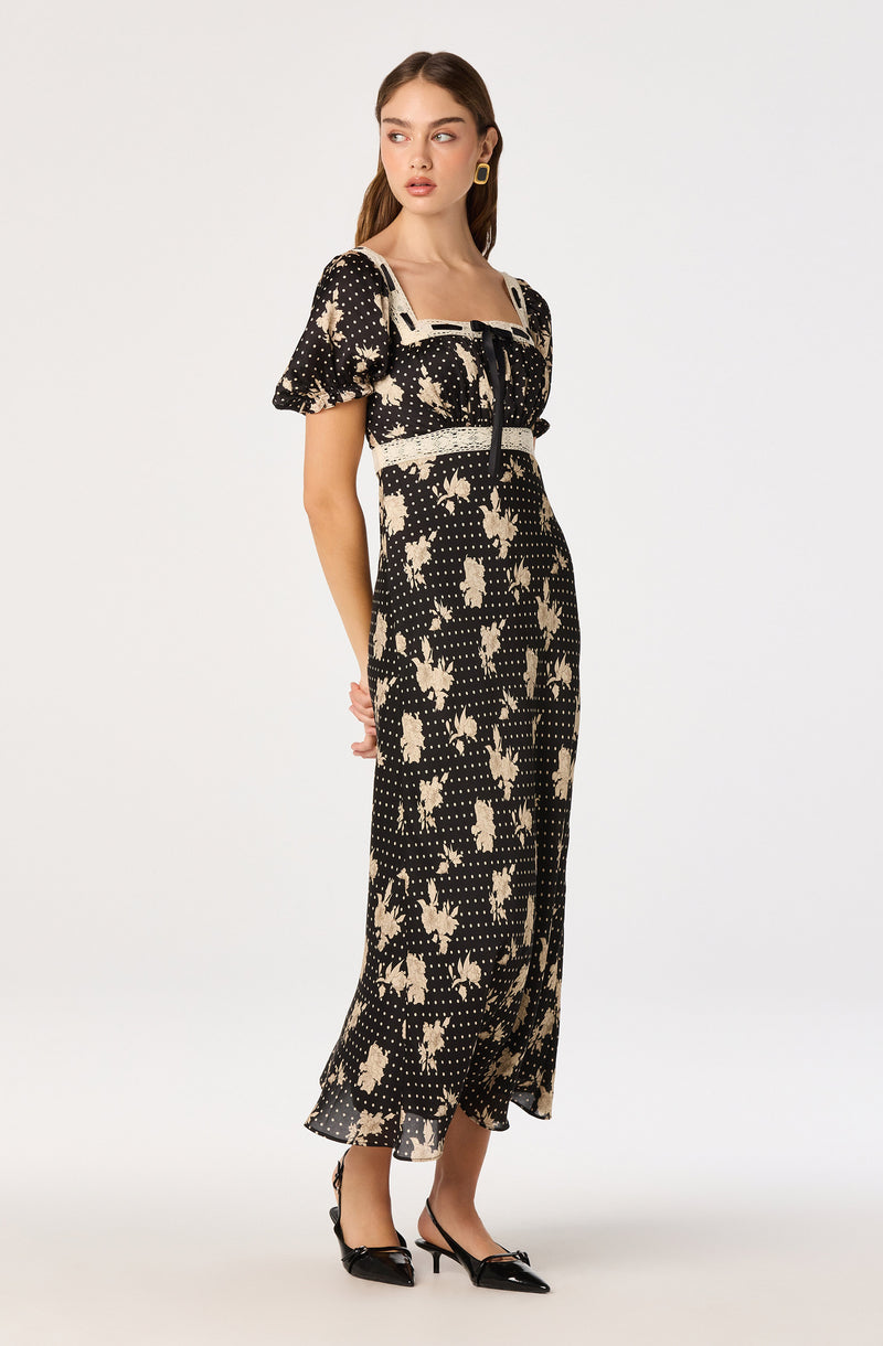 Woman wearing a black dress with cream floral and polka dot print, puff sleeves, and a square neckline.