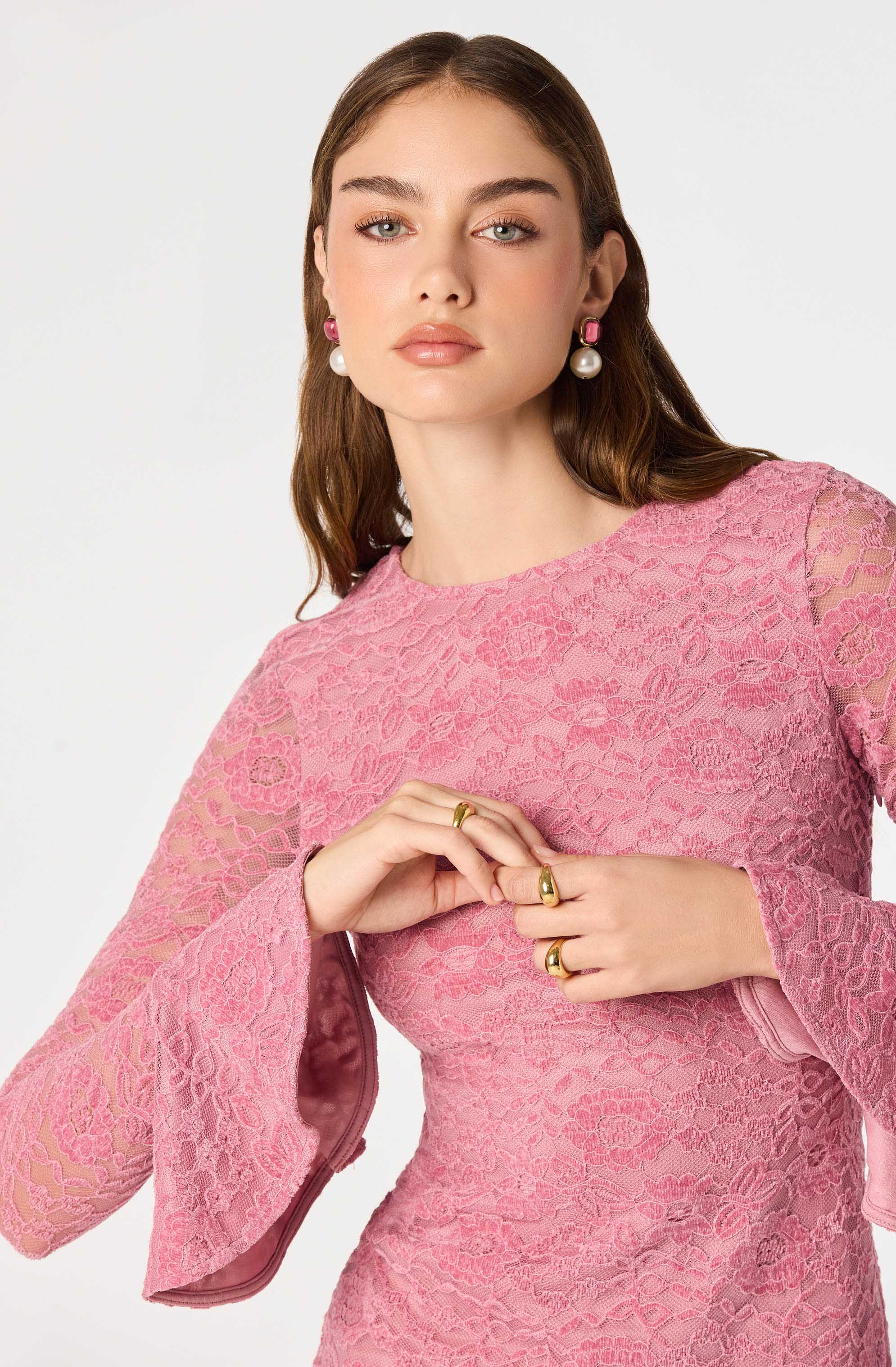 Woman in pink lace dress with bell sleeves, wearing pink and pearl earrings.