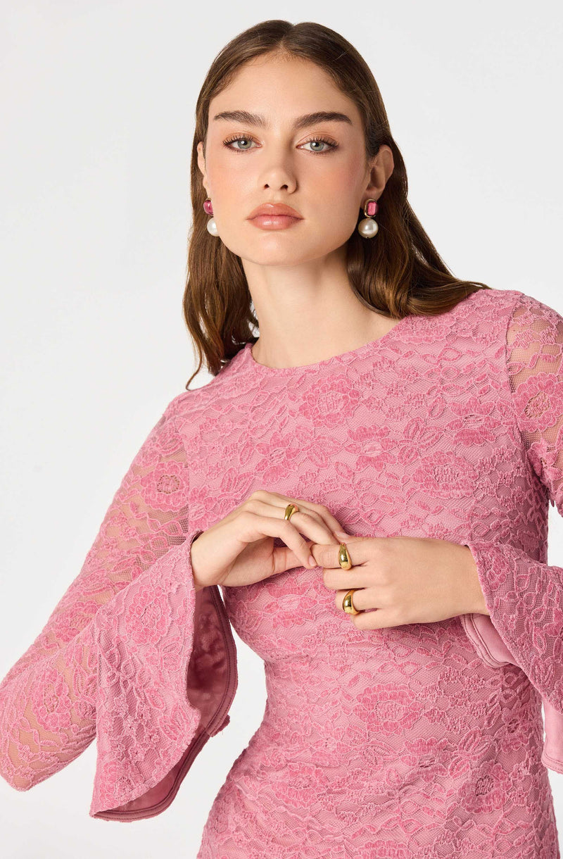 Woman in pink lace dress with bell sleeves, wearing pink and pearl earrings.