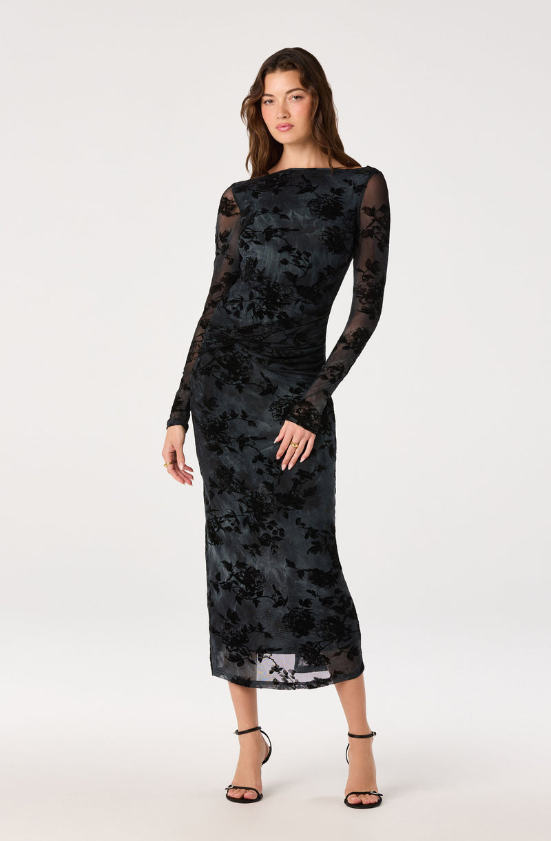 Woman in a black sheer long-sleeve dress with floral velvet pattern, paired with black high heels.