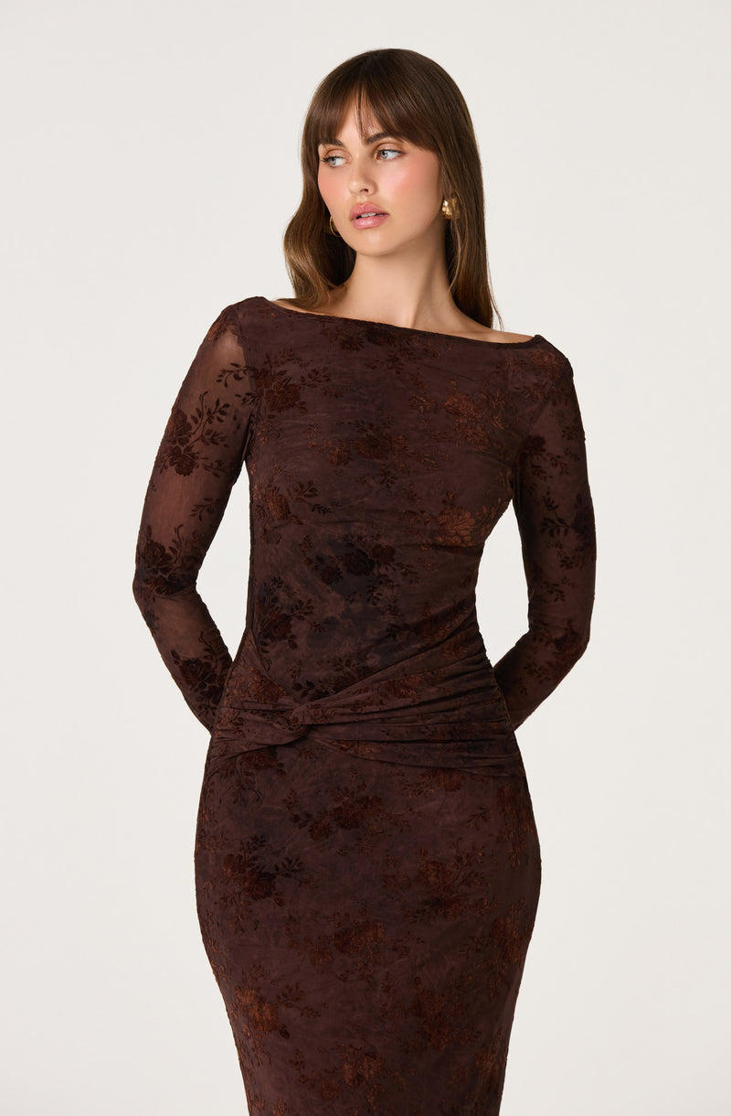 Woman in a long, sheer, brown lace dress with floral patterns and a knot detail at the waist.