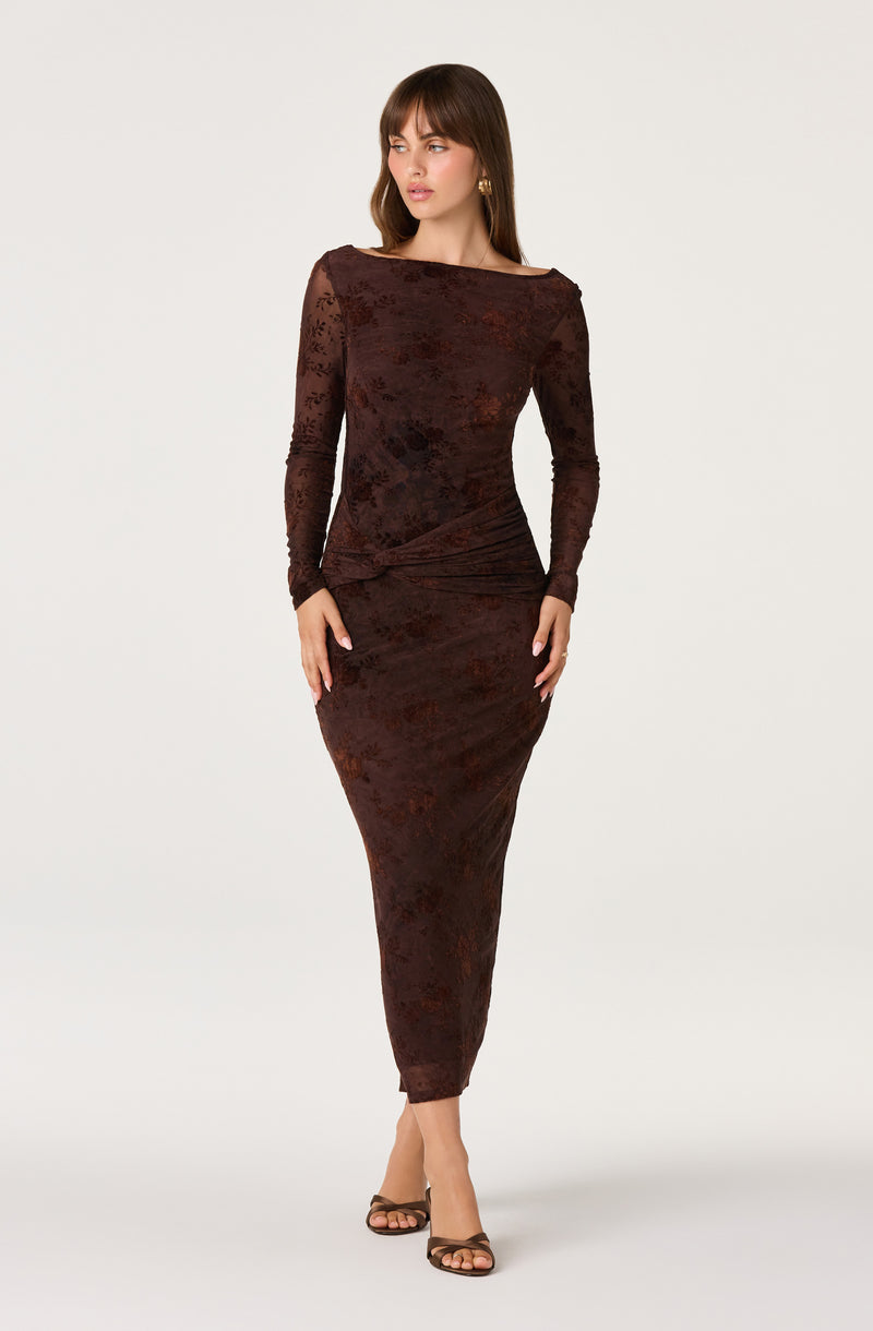 Woman wearing a long, fitted, dark floral lace dress with long sleeves and a boat neckline, paired with open-toe heels.