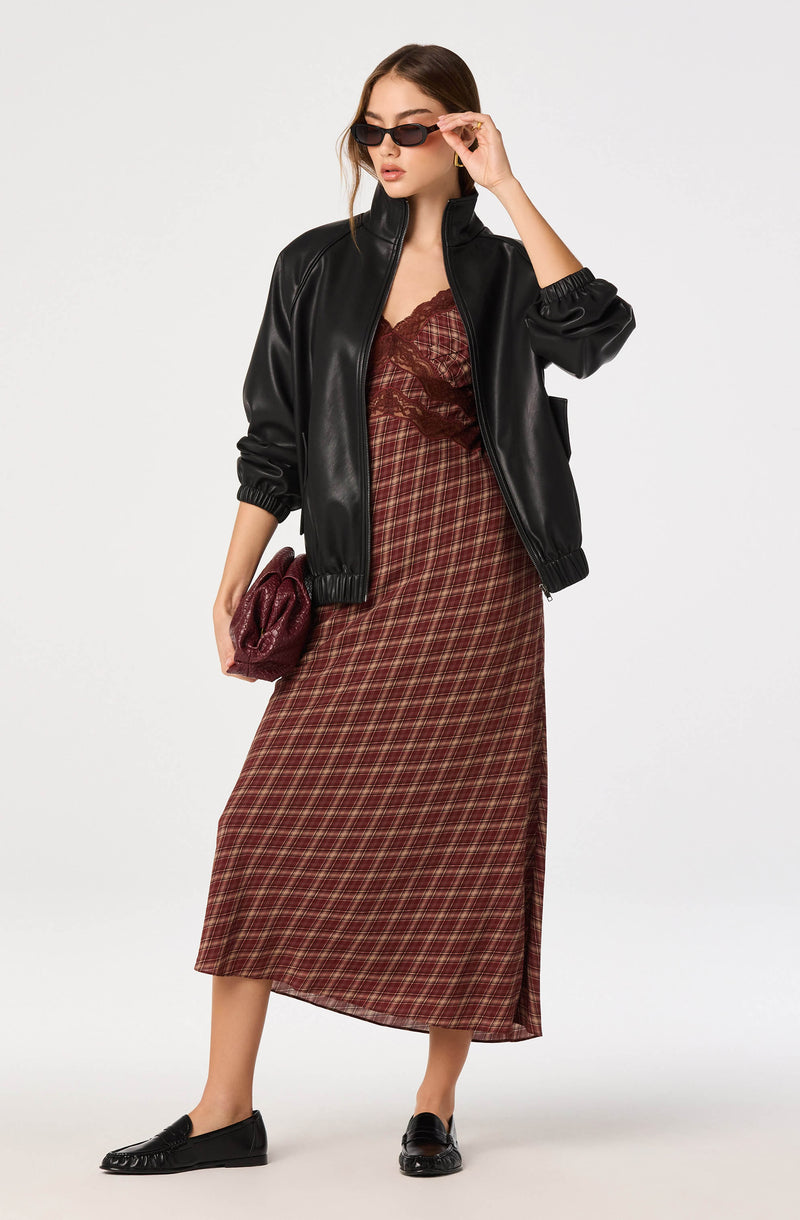 Woman wearing a black leather jacket, maroon plaid dress, black loafers, and dark sunglasses, holding a red clutch.