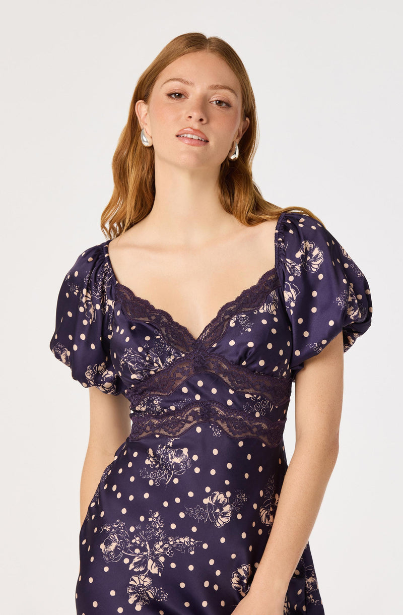 Young woman in a navy dress with puffed sleeves, floral and polka dot pattern, lace details.