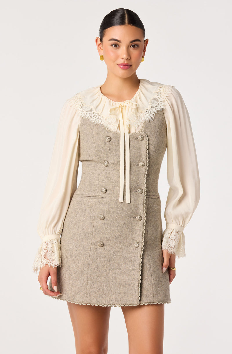 Woman wearing a beige dress with lace collar and cuffs, and a cream blouse with long sleeves.