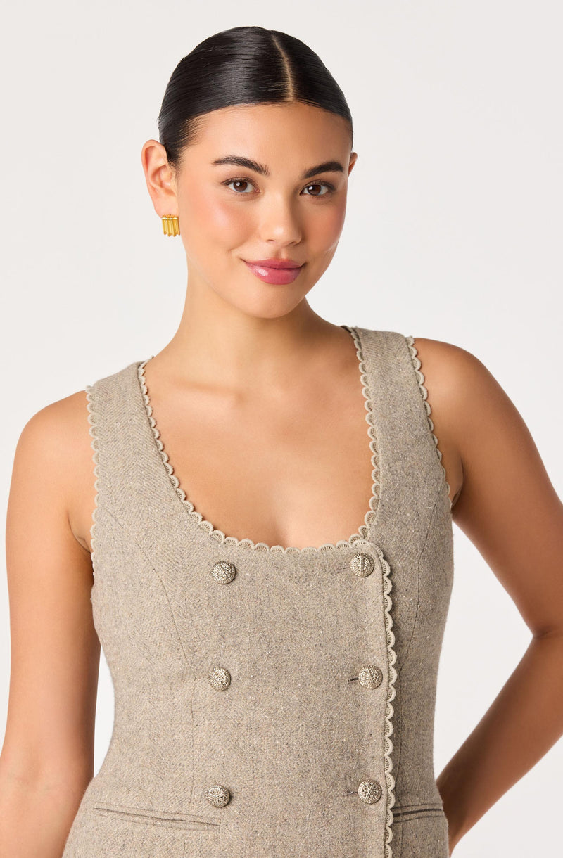 Woman wearing a beige tailored sleeveless dress with decorative buttons and scalloped edges.