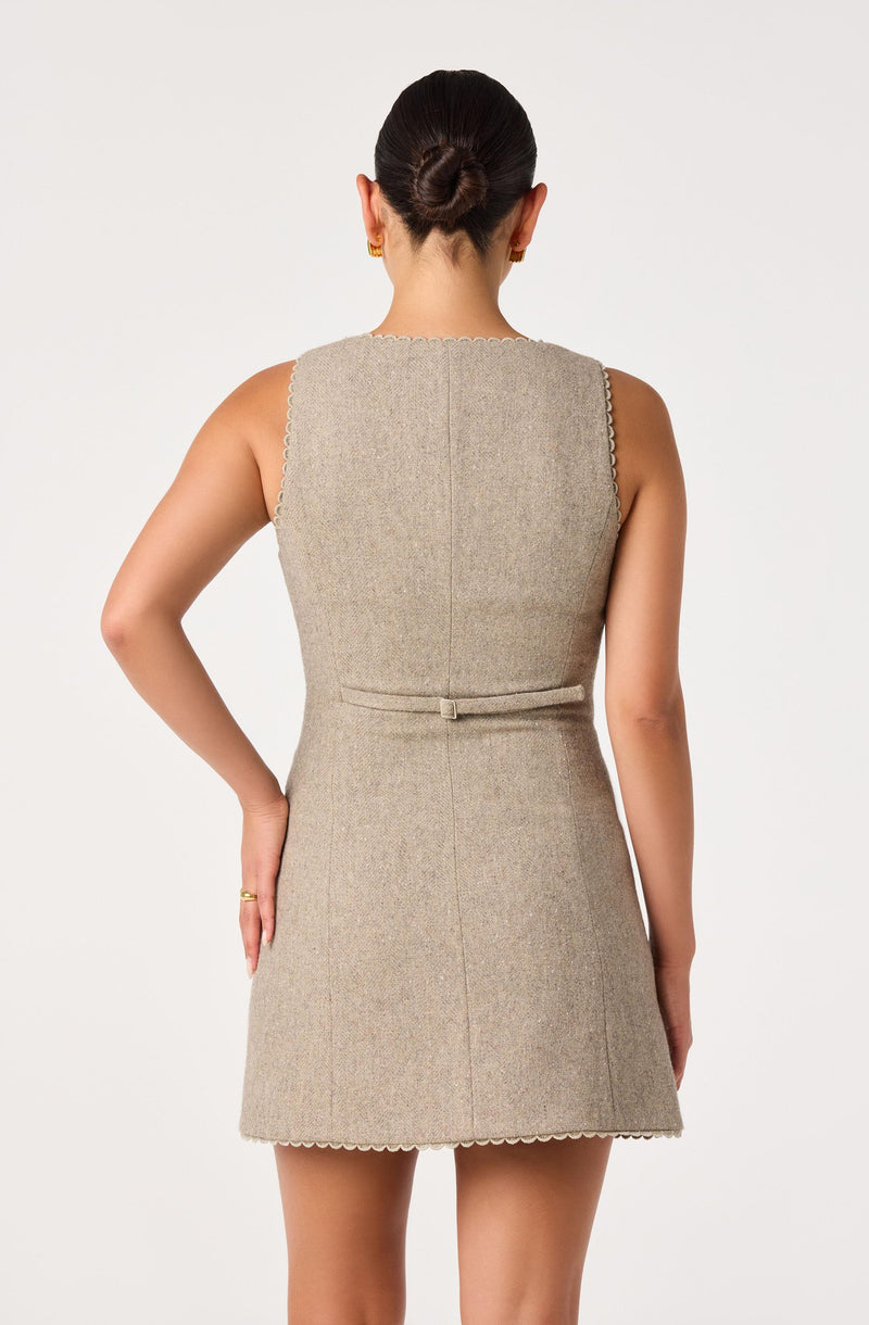 Back view of a woman wearing a sleeveless, beige dress with a textured knit pattern and decorative scalloped edges.