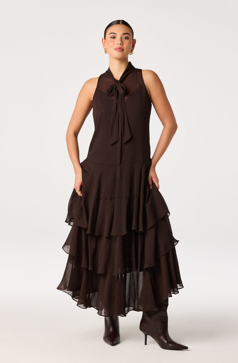 Woman in a sleeveless brown dress with a bow tie neckline and layered ruffle skirt, paired with brown heeled boots.