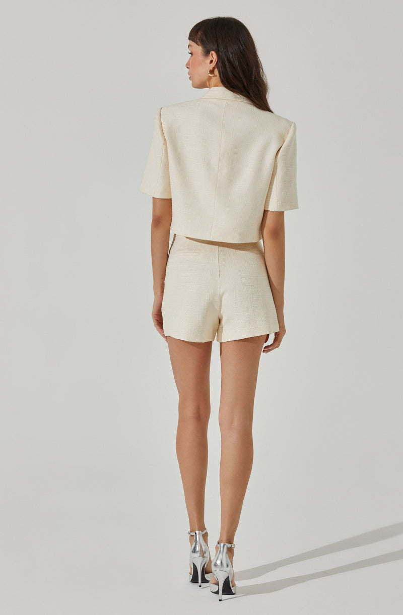 A woman is wearing a cream-colored, textured, short-sleeved blazer with structured shoulders and matching high-waisted shorts. She is also wearing shiny silver high-heeled shoes with ankle straps.