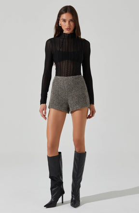 A woman is wearing a black, long-sleeved, semi-sheer, high-neck top with vertical stripes. She pairs it with gray, high-waisted, tailored shorts with a textured fabric. She completes the outfit with knee-high black leather boots with pointed toes and stiletto heels.