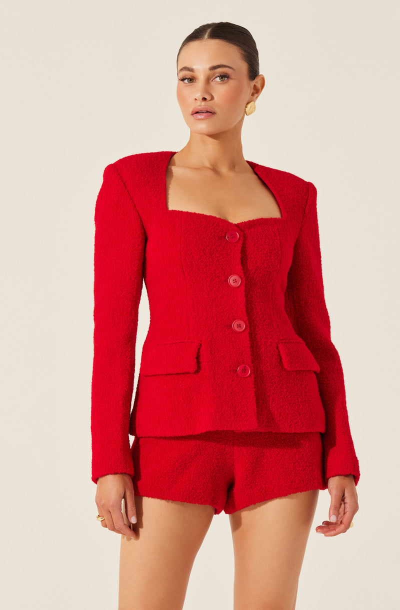A woman is wearing a bright red textured suit consisting of a fitted blazer with a square neckline, long sleeves, and four large buttons down the front. The blazer has two flap pockets on the front. She is also wearing matching red textured shorts. The outfit has a tailored, sophisticated style.