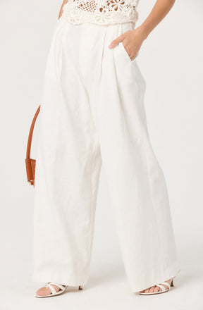 The image shows a person wearing high-waisted, wide-leg white pants with a relaxed fit. The pants have pleats at the waist and side pockets. The person is also wearing a white, sleeveless, crochet top with floral patterns. They have their right hand in their pocket and are wearing white strappy high-heeled sandals. A brown handbag with a thin strap is partially visible hanging on the person's left side.
