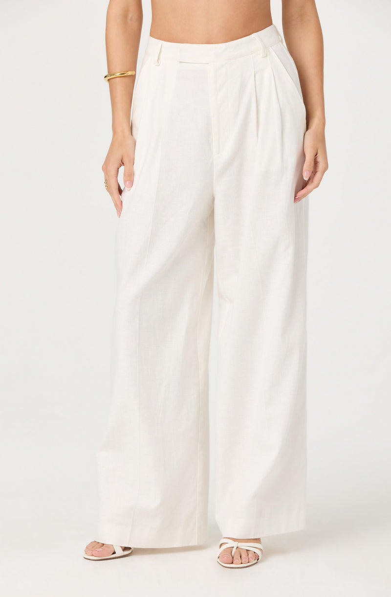 A person is wearing high-waisted, wide-leg white trousers made from a light, flowy fabric. The trousers have a tailored waistband with belt loops and pleats at the front. The person is also wearing a gold bracelet on their right arm and white strappy sandals.