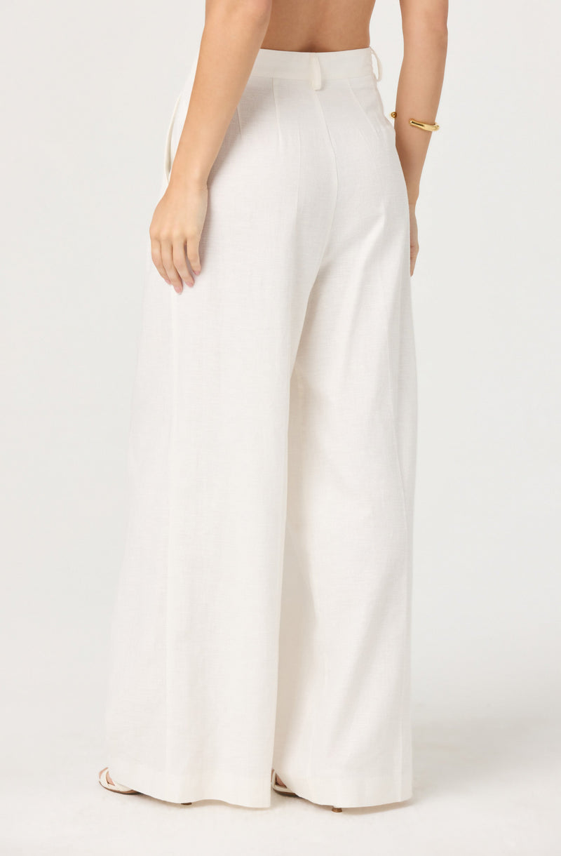 A person is wearing high-waisted, wide-leg white trousers made of a light, textured fabric. The trousers have a clean waistband with no visible belt, and they extend down to cover the shoes. The person is accessorizing with a simple gold bracelet on their right wrist.