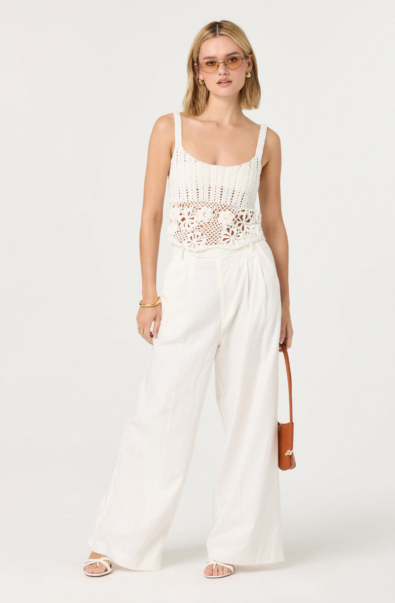 A woman is wearing a white crochet tank top with flower and geometric patterns and wide straps. She is also wearing high-waisted, wide-leg white trousers. She accessorizes with gold hoop earrings, a gold bracelet, and sunglasses with tinted lenses. She holds a small brown handbag with a strap in her right hand and wears white open-toe sandals.
