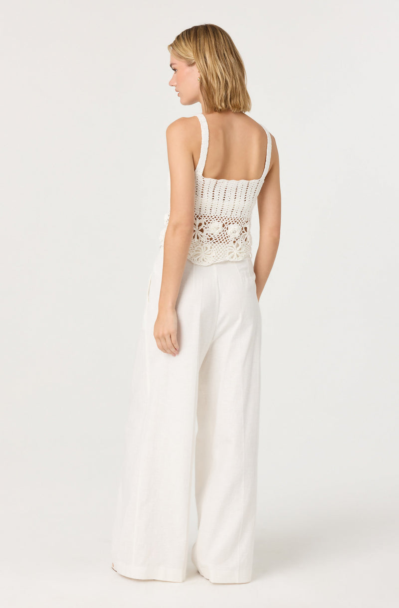 A woman is wearing a sleeveless, white, crocheted tank top with a fitted bodice and wide shoulder straps. She pairs it with high-waisted, wide-leg white trousers. The top features intricate openwork patterns, and the overall look is light and summery.