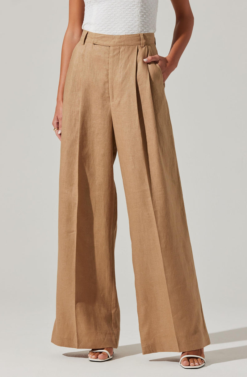 A woman is wearing high-waisted, wide-leg beige trousers with a pleat detail at the front and a matching fabric belt. The trousers have belt loops and a relaxed, flowing fit. She is also wearing a white textured sleeveless top.