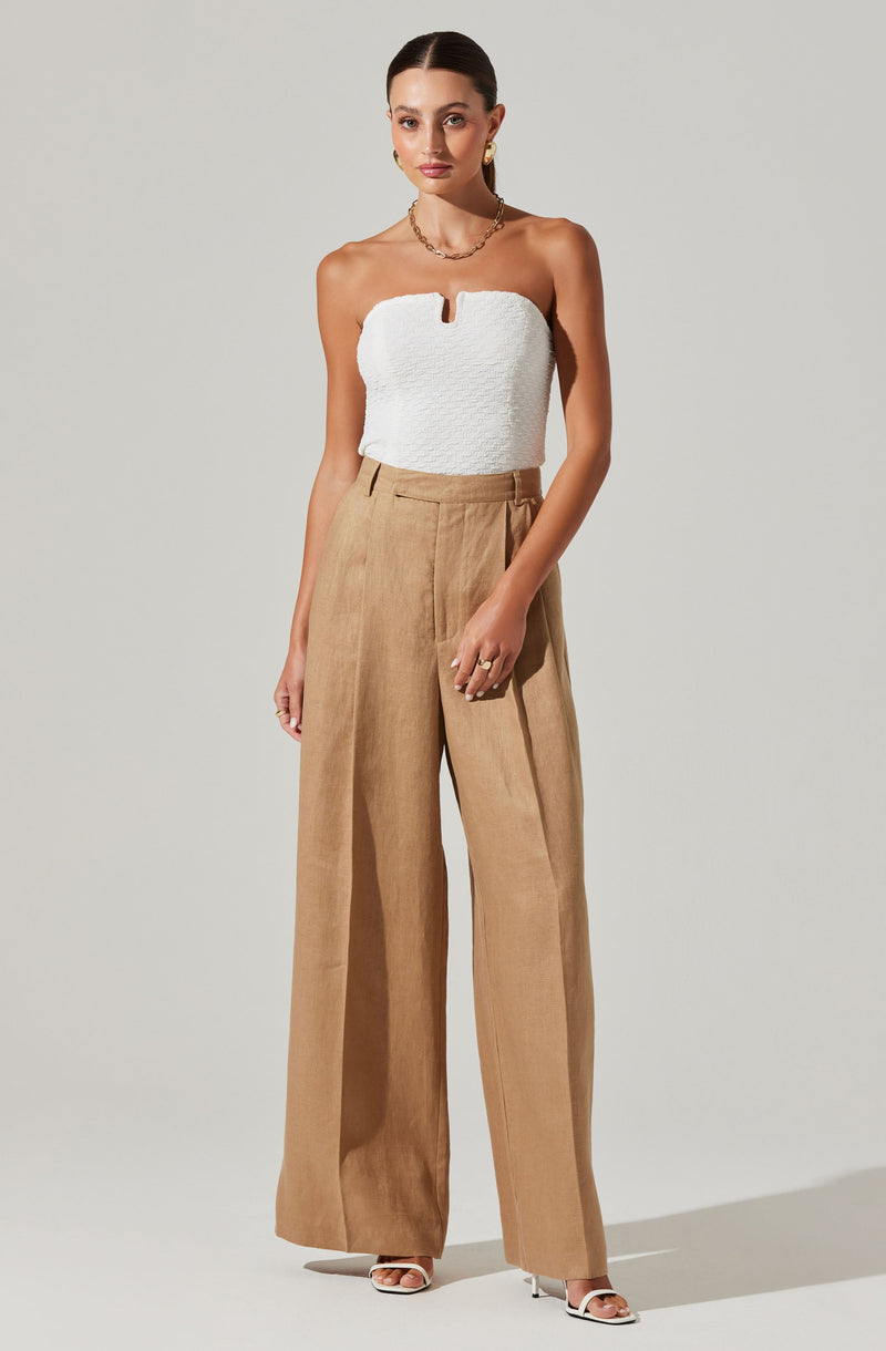 A woman is wearing a strapless white top with a textured fabric and a small notch in the center at the neckline. She is also wearing high-waisted, wide-leg tan trousers that have a tailored fit and a pleated front. She accessorizes with gold earrings, a gold chain necklace, and rings on her fingers.
